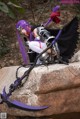 A woman with purple hair sitting on a rock holding a sword.