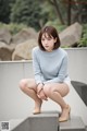 A woman in a blue sweater and white shorts sitting on a concrete ledge.